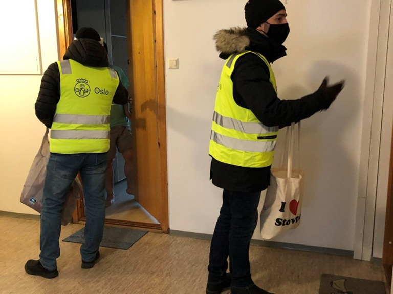 Dør til dør-aksjon med en mann iført refleksvest med ryggen til og en med munnbind informerer innendørs i en oppgang